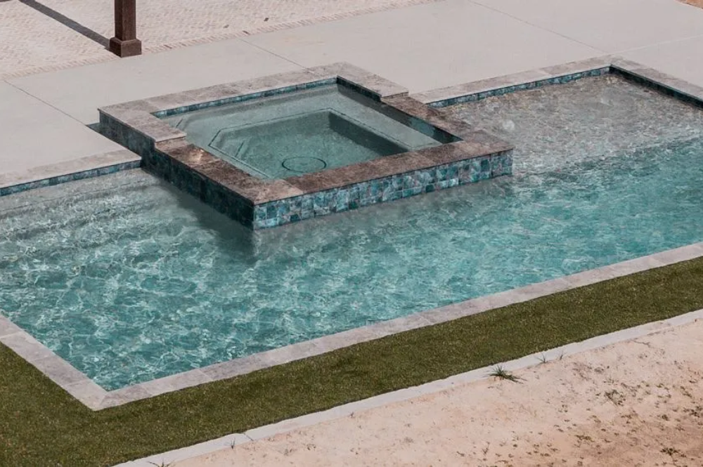 Aerial view of a modern swimming pool and spa, featuring clear blue water, stone borders, and surrounding concrete pavement and grass